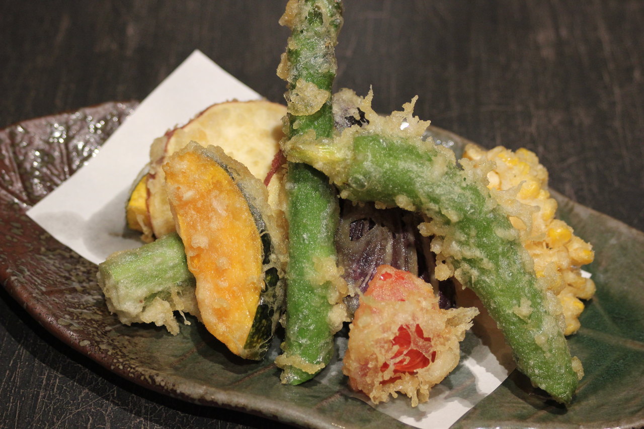 几朵炸至金黄的小黄瓜花天妇罗整齐摆放在黑色石板盘中,花瓣形态清晰,外层酥皮泛着光泽,旁边配有小型陶瓷碟盛放棕色天妇罗蘸汁,背景虚化的日式料理环境 小黄瓜花天妇罗摆盘展示