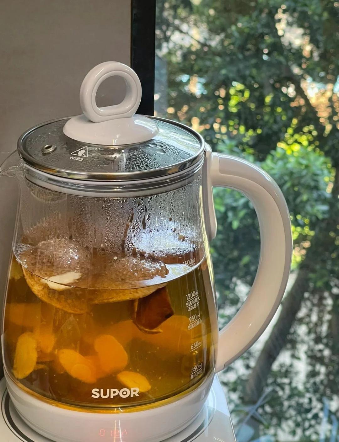 透明玻璃锅中正在熬煮沙田柚皮,水中漂浮着切成条状的黄色柚皮和两片生姜,蒸汽袅袅升起,灶台为现代厨房环境,旁边摆放着干净的瓷杯和木勺,展现家庭自制健康饮品的真实场景 沙田柚皮煮水过程图