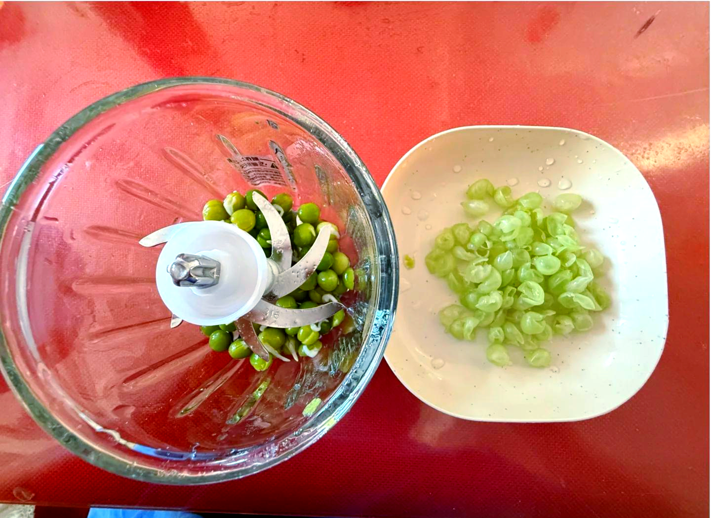 厨房操作台场景：一只手正在用剪刀修剪豌豆尖的卷须，旁边放着小碗装的小苏打和醋，背景有沥水篮和清水盆