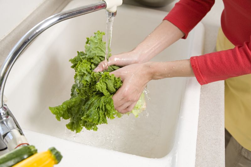 一位家庭主妇在厨房水槽前用手轻轻冲洗一把油麦菜,水流清澈,蔬菜鲜绿,旁边放着菜篮和砧板,背景温馨整洁,体现日常生活中的健康饮食场景 家庭厨房中正在清洗油麦菜的场景