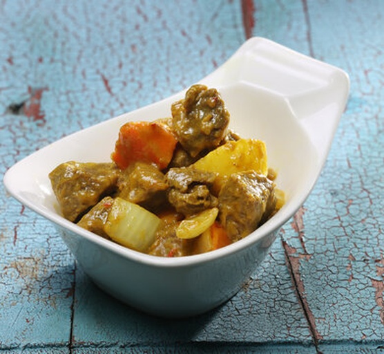 一碗盛满的黄色咖喱菜肴,配有土豆、胡萝卜和鸡肉块,表面撒有少许现磨黑胡椒粉,旁边放置一把木勺,背景虚化,突出食物的诱人色泽与质感 成品咖喱菜肴特写