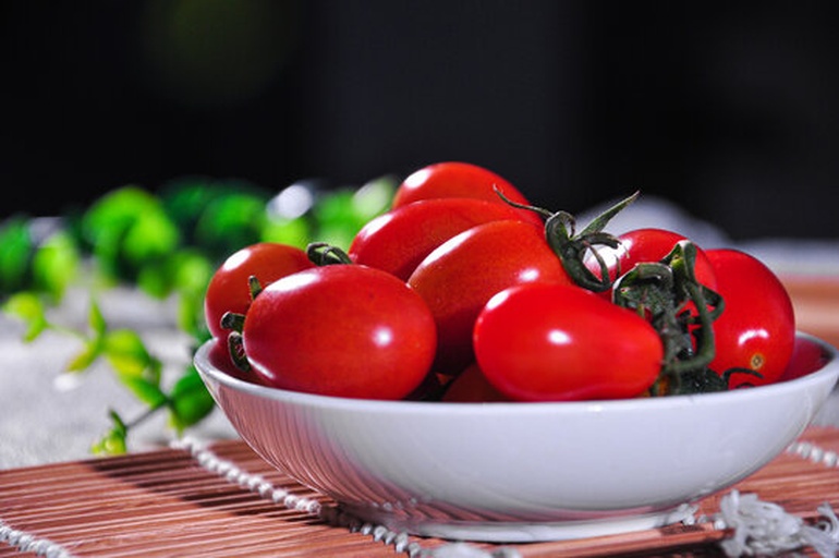 一位笑容温暖的中年女性正将新鲜红润的樱桃番茄倒入白瓷碗中，背景是阳光洒落的现代厨房台面，案板上散落着几枚饱满多汁的牛心番茄和一把翠绿罗勒，自然光线下番茄表皮泛着水润光泽