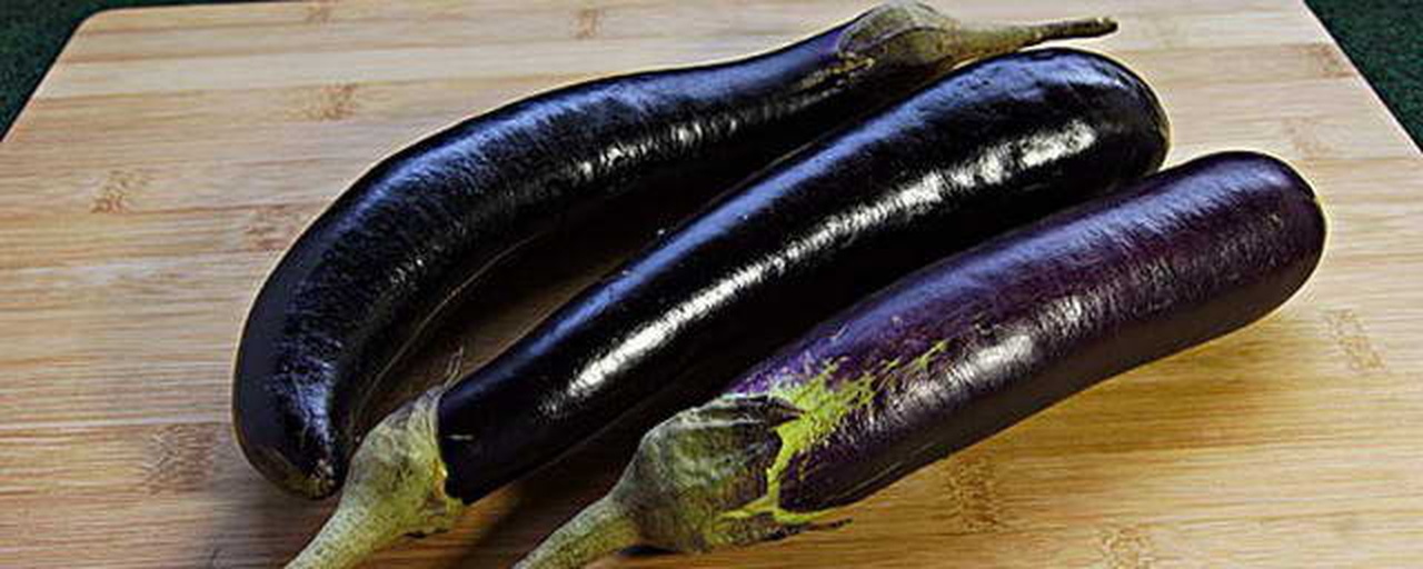 一位家庭主妇在厨房砧板上用刀切开一个紫黑色长茄子,露出洁白蓬松的果肉和隐约可见的种子,旁边摆放着完整的带萼片茄子,背景为现代厨房环境,突出生活化场景 家庭厨房中切开茄子的场景图