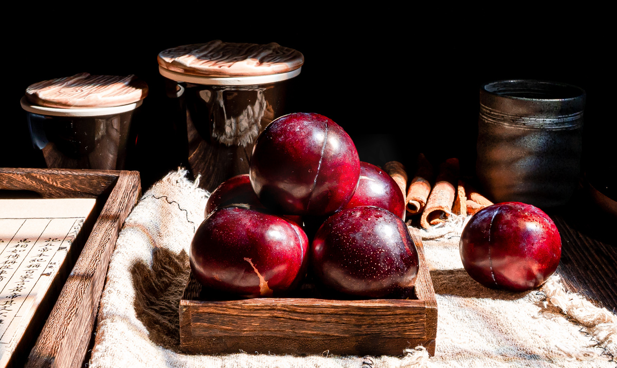 一张木质餐桌上摆放着白色瓷盘,盘中整齐码放着6颗完整的蜂糖李,旁边有一小碗去核切块的果肉,搭配薄荷叶点缀,背景虚化的玻璃杯中盛有淡黄色果汁,整体呈现高级简约的美食摄影风格 蜂糖李餐桌摆盘示意图