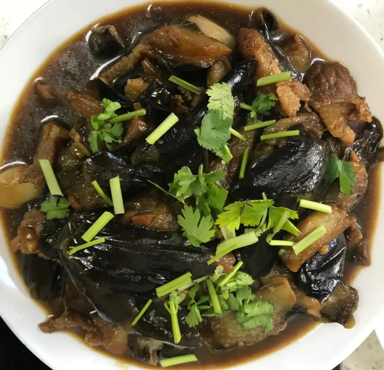 一盘刚出锅的家常炖茄子,色泽红亮,酱汁浓稠,茄子块完整饱满,表面泛着油光,顶部撒有翠绿葱花,盛放在白色瓷盘中,背景为木质餐桌 成品家常炖茄子俯拍图