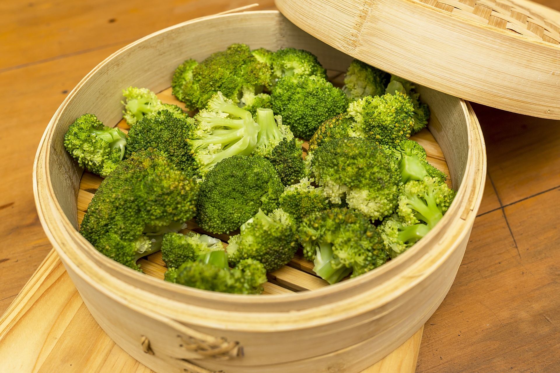 一个透明锅盖的蒸锅内正在加热松花菜,可见蒸汽升腾,花球呈淡黄色,置于竹制蒸笼上,旁边放置一个小碗装有橄榄油和切碎的蒜末,体现健康烹饪理念 厨房中正在清蒸松花菜的场景图