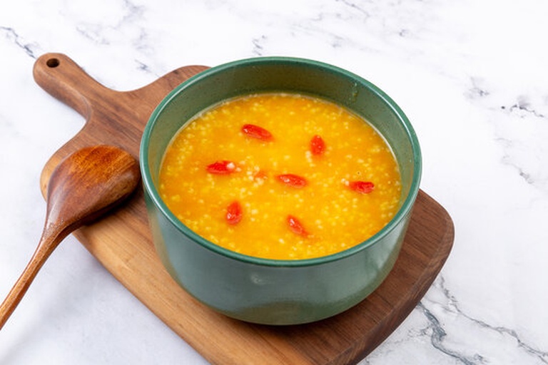 一口冒着热气的白色砂锅正在炉灶上慢火熬煮，锅盖微开，可见金黄色小米粥翻滚，旁边摆放着泡好的枸杞小碟