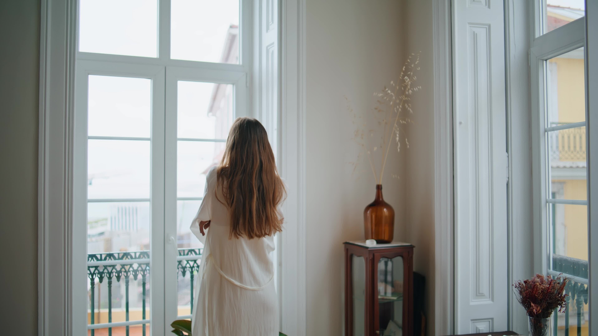 一位30岁左右女性站在厨房窗边,身穿浅色棉麻围裙,双手捧着透明玻璃杯,杯中为深褐色略带绿意的液体,阳光透过窗户洒在杯子上,背景有绿植与木质橱柜 女性手持玻璃杯饮用黑豆车前草水