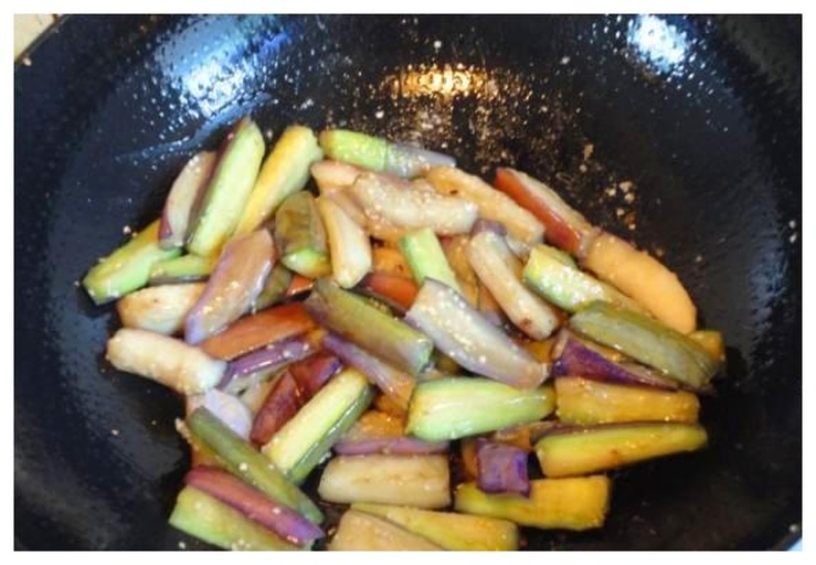 一位厨师正在灶台前翻炒锅中的茄子,锅内茄子呈金黄色,正在加入深色酱汁,周围摆放着葱姜蒜、酱油瓶、碗碟等厨具,环境整洁明亮,展现家庭厨房的真实操作画面 厨房中正在烹饪红烧茄子的过程场景