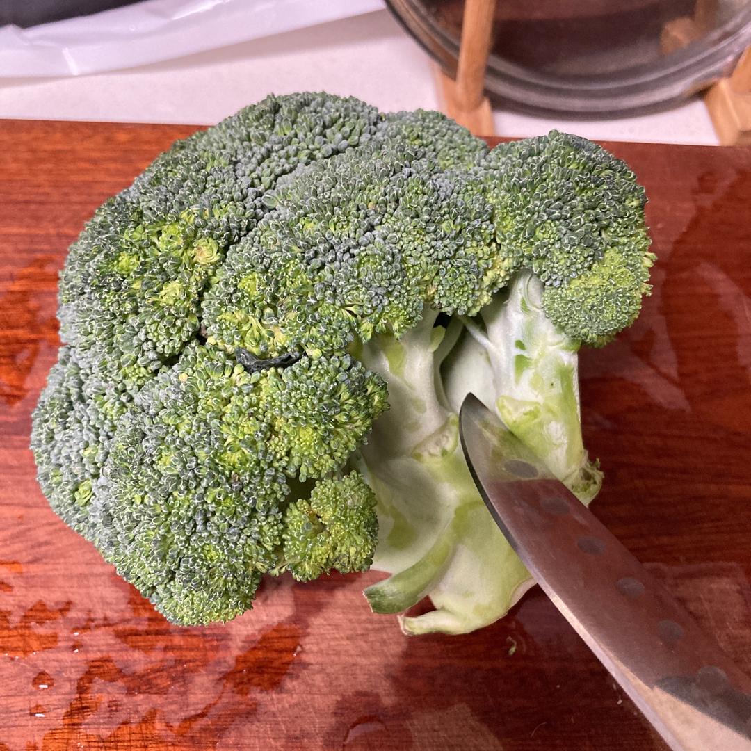 一位家庭主妇在厨房操作台上将西兰花切成小朵,旁边有干净的砧板、刀具和一碗清水,背景可见冰箱门打开,整体氛围温馨整洁,体现日常食材处理情境 家庭厨房中正在处理西兰花的场景