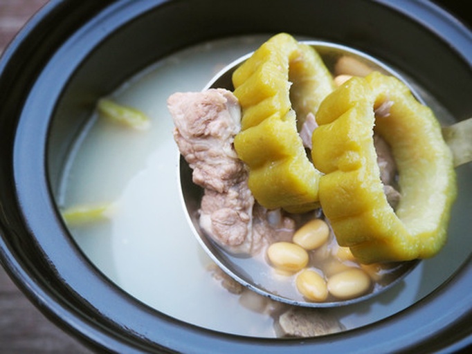 一口不锈钢汤锅正在炉灶上加热,锅内清水翻滚,漂浮着几段切好的苦瓜根和姜片,旁边摆放着漏勺和一碗生排骨 苦瓜根在锅中焯水过程