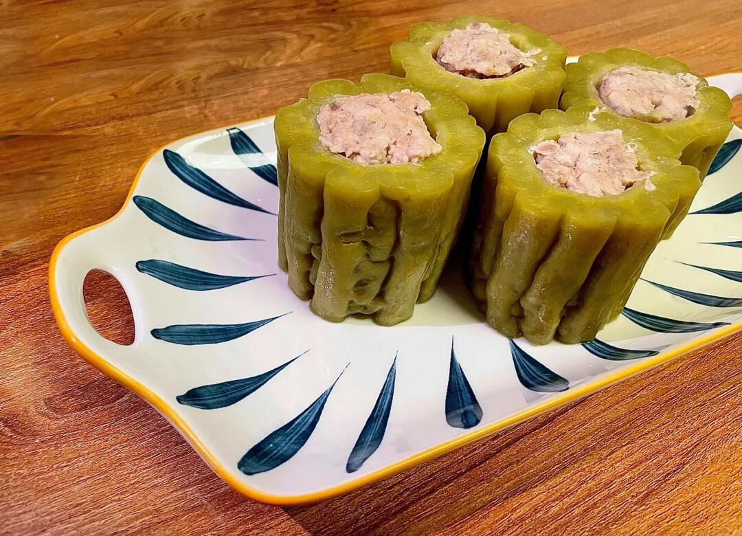浅色木桌上摆放一盘苦瓜酿肉,苦瓜切成圆环状,中间填充褐色肉馅,表面微焦泛光,周围点缀香菜叶,整体呈现中式家常菜肴风格 苦瓜酿肉成品摆盘图