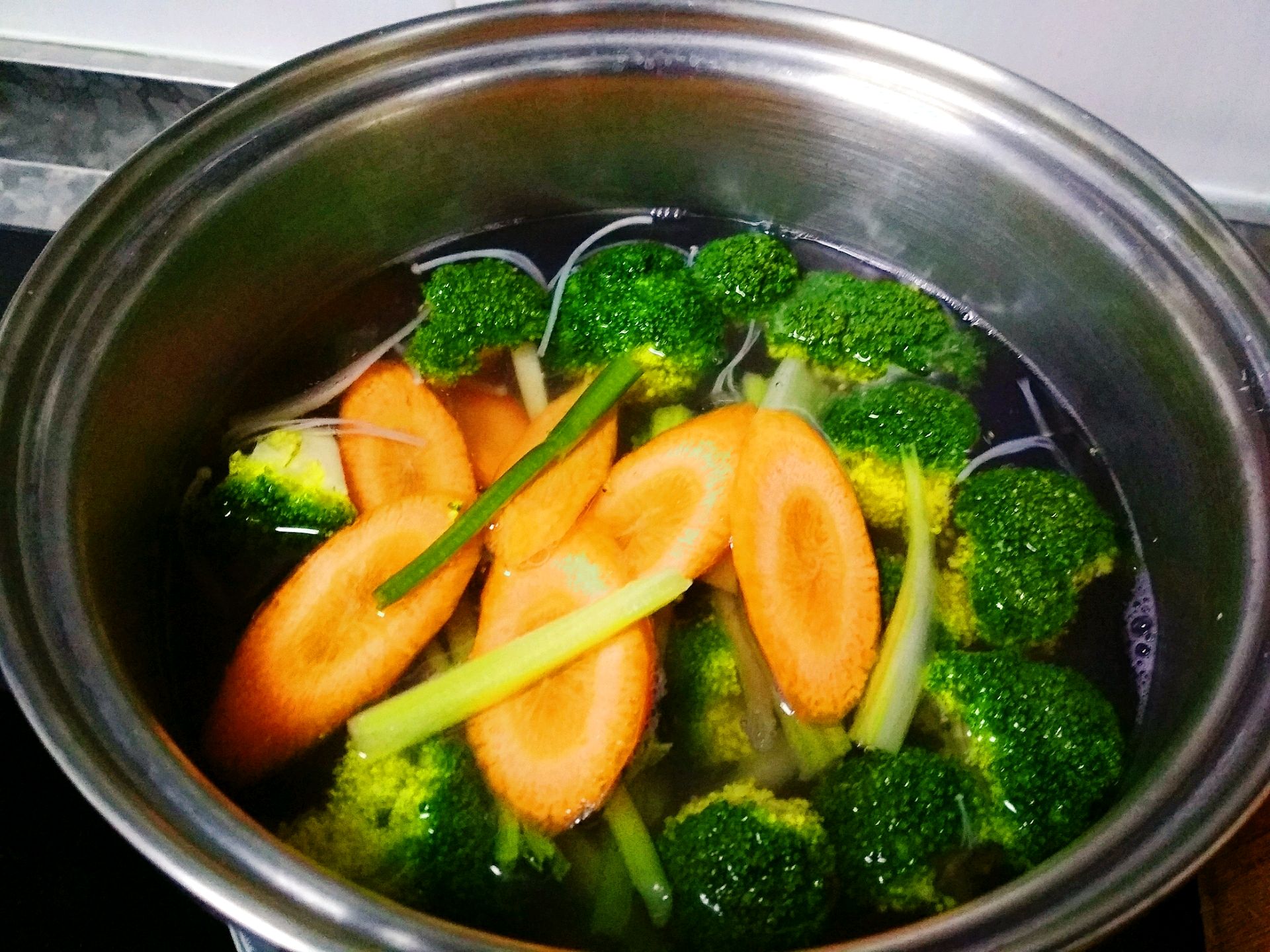 一口白色陶瓷锅中炖煮着多种蔬菜,包括发黄的西兰花、胡萝卜片和土豆块,汤面微微冒泡,热气腾腾,展现出利用稍老西兰花制作家常菜肴的健康方式 一锅正在炖煮的蔬菜汤,其中包含黄色和绿色的西兰花块