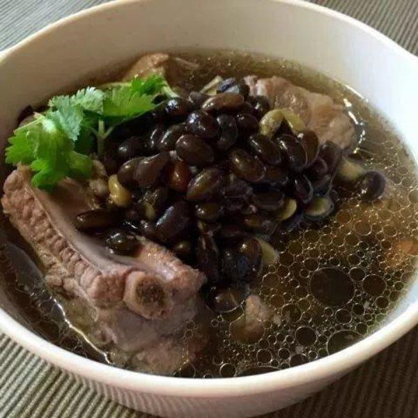 一锅热气腾腾的黑豆炖猪骨汤,深褐色汤汁浓郁,黑豆饱满,搭配几片胡萝卜和葱花点缀,盛放在白色瓷碗中,背景为厨房木质台面 黑豆料理成品图