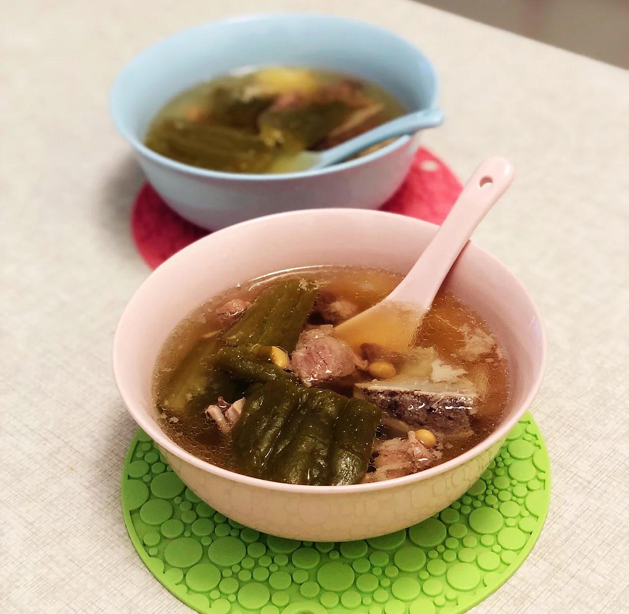 一碗盛满的苦瓜骨头汤,汤色呈乳白色略带淡黄,表面漂浮着几片翠绿的苦瓜、完整的黄豆和蜜枣,旁边配有瓷勺,背景虚化显示木质餐桌和竹筷,整体呈现温馨家常氛围 成品苦瓜骨头汤特写