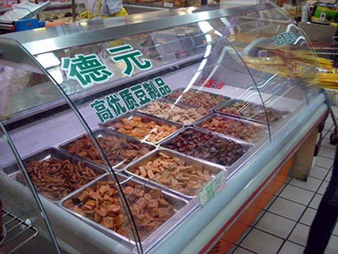 一个现代超市冷柜货架,陈列多种豆制品:透明包装的嫩豆腐、塑料盒装的北豆腐、真空袋装的豆腐干、小杯装纳豆、纸盒装无糖豆浆,标签清晰可见,展示市售豆制品多样性 超市货架上的豆制品专区