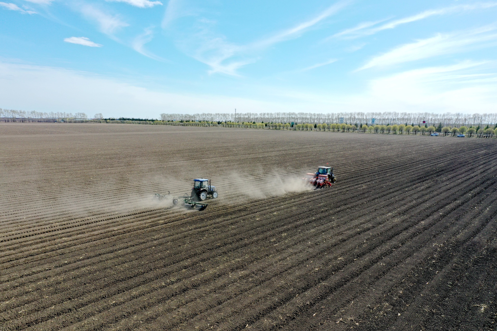 一台拖拉机牵引着油菜直播机在平整的农田中作业,土壤翻松均匀,播种机正在精准下种,远处有农民在查看播种深度,背景为初春湿润的田野环境 油菜田机械化播种现场