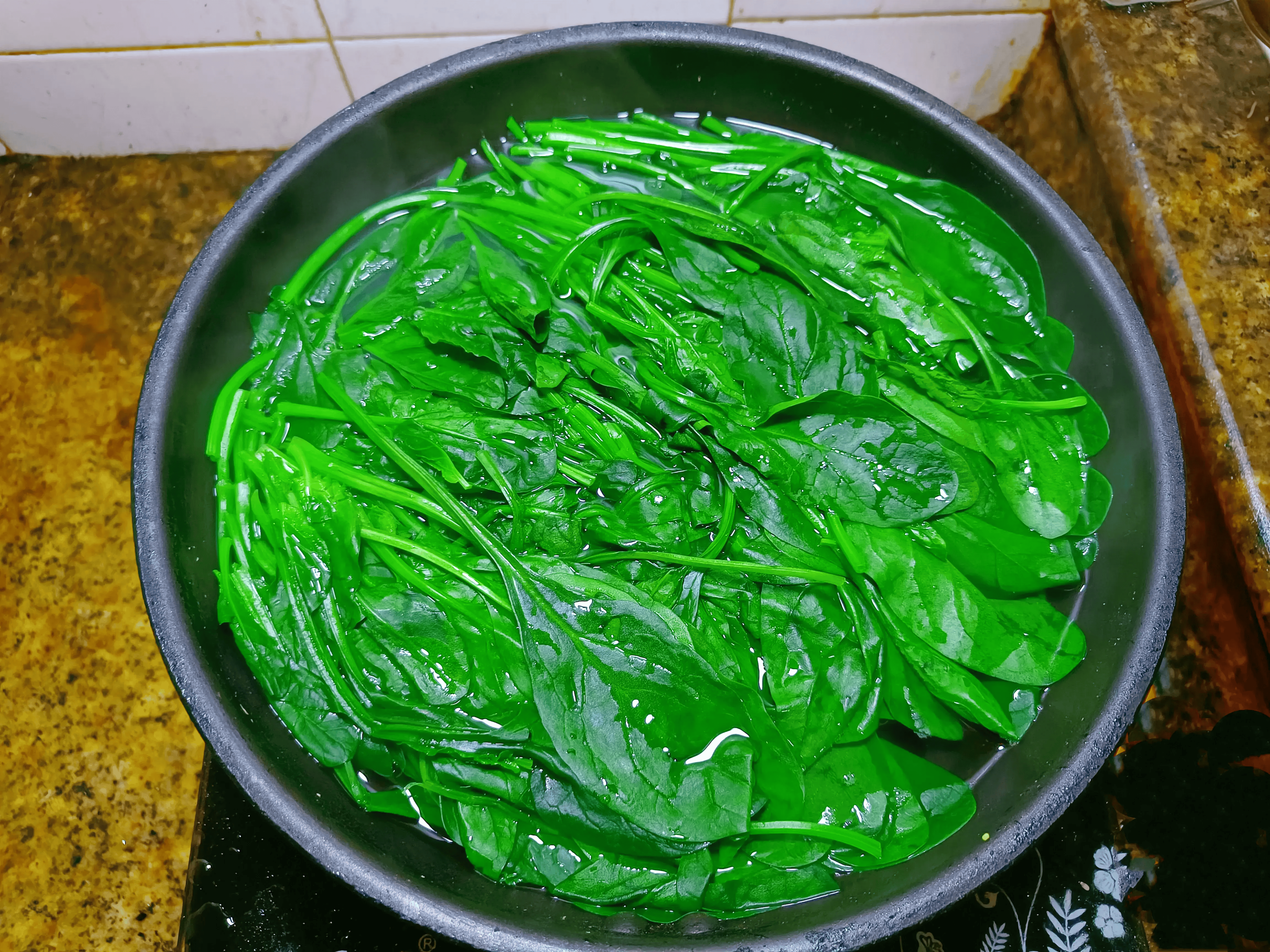 一位女性在明亮的现代厨房中,正在水槽里用手清洗一把深绿色的新鲜菠菜,水流清澈,菠菜叶片完整无损,旁边放着砧板和刀具,背景可见冰箱和橱柜 家庭主妇在厨房清洗与处理新鲜菠菜的过程