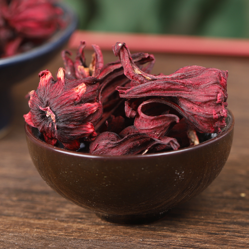 一株生长旺盛的洛神花植株,背景为热带农田,前景展示刚采摘下来的完整花萼,呈深紫红色,表面有细微绒毛,质地厚实多汁,旁边摆放白色瓷盘盛放干燥后的洛神花干品 洛神花植株与新鲜采摘的紫红色花萼特写