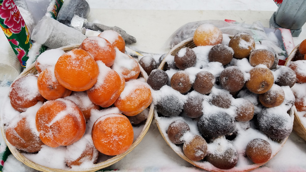 一家三代人围坐在东北火炕上,桌上摆着几颗冻柿子和一杯热水,老人正在教孩子如何用 straw 吸食冻柿子,窗外飘着雪花,充满温馨的家庭氛围 一家人围坐火炉旁享用冻柿子