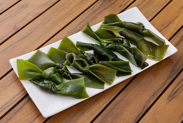 一盘精致的海带包饭摆放在木质餐盘中,每根海带卷整齐切段,露出内部饱满的米饭,表面微微泛光,旁边点缀着胡萝卜丝和芝麻,背景为浅色厨房台面,光线柔和自然 海带包饭成品特写