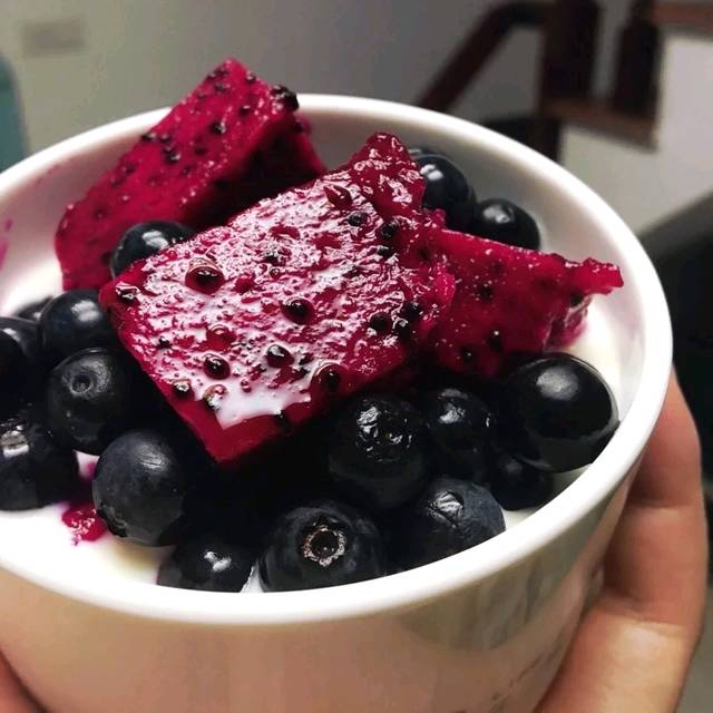 火龙果与蓝莓并排摆放于婴儿餐盘中，左侧为切开的白心火龙果果肉，右侧为新鲜蓝莓颗粒，中间用硅胶婴儿勺轻压成微碎状态，背景为浅木纹辅食台，体现安全、洁净、适龄的喂养场景