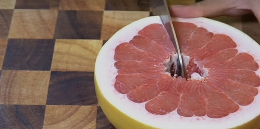 一位厨师正在砧板上使用水果刀为红肉葡萄柚削去外皮和白色海绵层,随后用刀尖沿果瓣边缘精细切割,分离出完整的半透明红色果肉瓣,背景为木质厨房台面,光线柔和自然 葡萄柚去皮取瓣过程