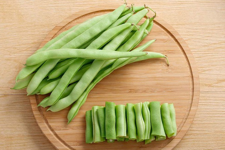新鲜绿色的长条豆角整齐摆放在白色砧板上,左侧为未处理的整根豆角,右侧为已切段并去除筋络的豆角段,背景有剪刀和菜刀工具 豆角处理前后对比图
