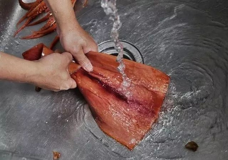 一位家庭主妇正在厨房的水槽边清洗笔管鱿鱼,案板上有几只已去内脏的鱿鱼,水珠飞溅,旁边放着生姜、葱段和料酒,准备用于后续烹饪 家庭厨房中清洗笔管鱿鱼的过程