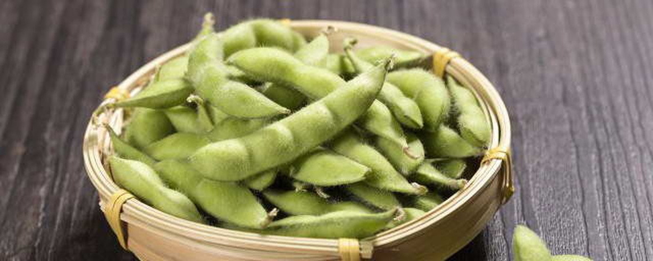 一位中年女性坐在阳台小凳上手工剥毛豆,面前放着装满带荚毛豆的竹筐和一只碗中已剥好的翠绿毛豆米,阳光洒落,体现温馨生活气息 家庭剥毛豆场景图