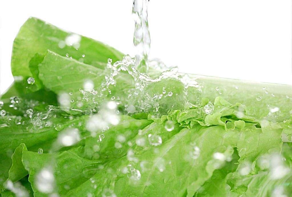 一盆新鲜的生菜正在透明玻璃盆中用流动清水浸泡,水中有细小气泡上升,背景为厨房台面,旁边放有滤网和干净毛巾,光线自然明亮 生菜在清水盆中浸泡清洗的场景