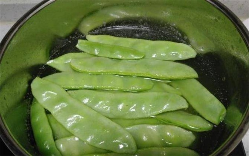 一位厨师正在灶台前翻炒锅中的扁豆,扁豆呈翠绿色,伴有蒜片和红椒丝,火焰适中,锅具为中式铁锅,背景为整洁厨房环境 家庭炒制扁豆过程