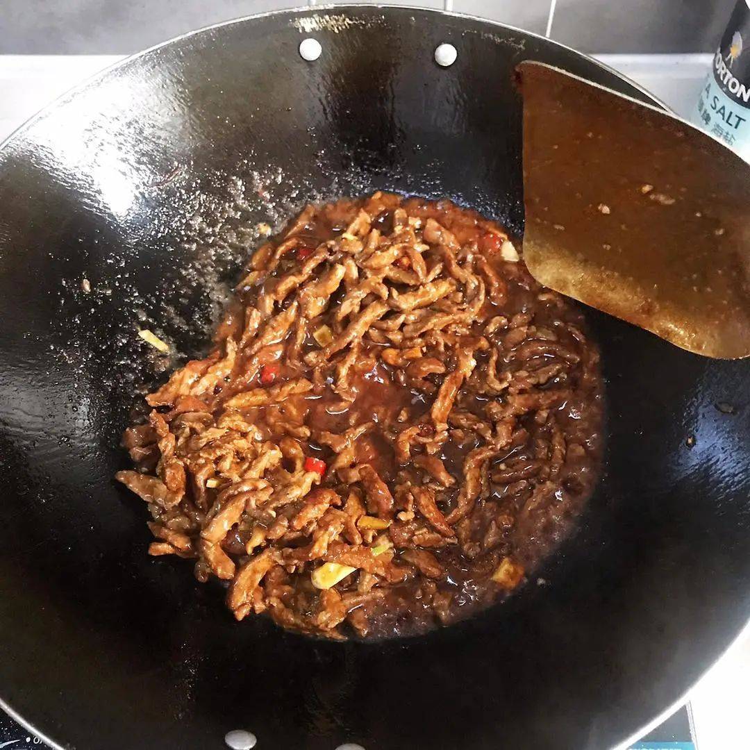 一位家庭主妇在明亮的现代厨房中正在用铁锅翻炒红菜苔,锅中菜苔呈紫红色,伴有蒜片和少量腊肉,热气腾腾,灶台旁摆放着切好的食材和调料瓶,背景为整洁的橱柜和窗户,光线柔和,营造温馨的家常烹饪氛围 家庭厨房中正在烹饪红菜苔的场景