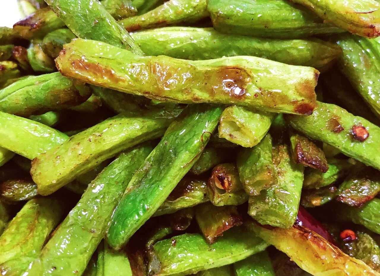 一盘新鲜绿色的四季豆分为两部分:左侧为未经烹煮的生四季豆,颜色鲜亮但质地坚硬;右侧为完全炒熟的四季豆,颜色略深呈墨绿,边缘微皱,表面有油亮光泽,明显软化。背景为厨房灶台,配有锅铲和炒锅 四季豆与生熟对比图