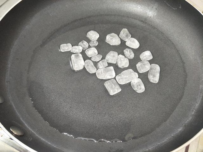 平底锅中正在炒制糖色,冰糖在油中融化,呈现出金黄色至琥珀色的浓稠液体,厨师手持锅铲持续搅拌,热气升腾,背景虚化的灶具显示正在使用中小火 炒糖色过程特写