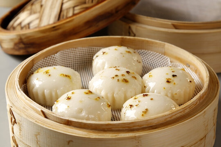 竹制蒸笼中整齐摆放着四个用绿色荷叶包裹成型的鸡腿,周围冒出白色蒸汽,旁边有一碗蒸出的鸡汤,背景为传统中式厨房灶台 香荷腊鸡腿蒸制过程图