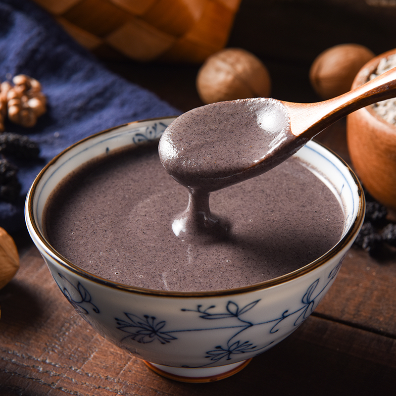 一位匠人使用传统青石磨盘手工研磨炒熟的黑芝麻,细腻的芝麻酱缓缓从磨缝流出,盛放在陶瓷碗中,旁边摆放着新鲜出炉的全麦馒头,展示传统中式早餐搭配场景 传统石磨研磨黑芝麻制作芝麻酱的过程