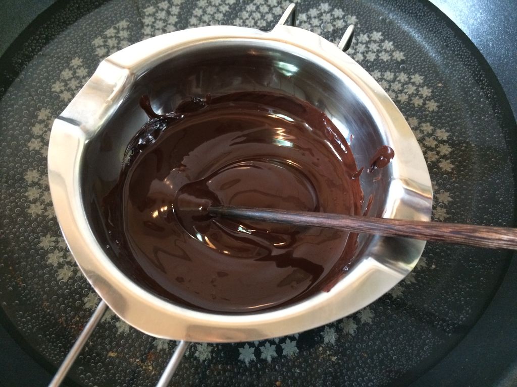 一个透明玻璃碗放置在盛有热水的不锈钢锅上,碗中黑色巧克力正在缓缓融化,一只手正用白色硅胶铲搅拌,水面蒸气轻微上升,画面清晰展示隔水加热的操作细节 巧克力融化过程