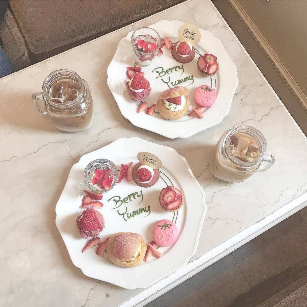 晶莹剔透的高脚杯中盛放着淡粉色草莓酒，背景为白色桌布与奶油色甜点拼盘，包含马卡龙、芝士蛋糕和新鲜浆果