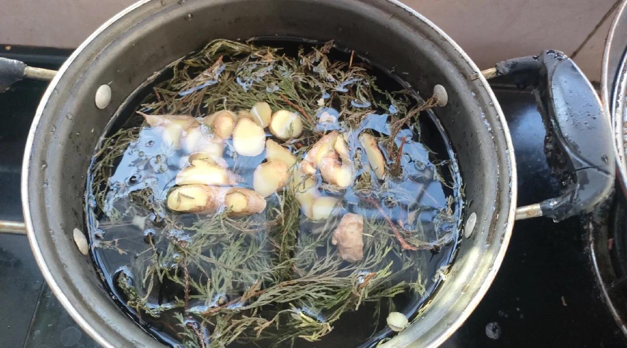 一位女性正在厨房用砂锅煎煮侧柏叶药材,锅边冒着蒸汽;旁边桌上放置一碗深褐色药液和一个木梳,展示准备用来洗头的过程 侧柏叶煎煮过程与洗头示意