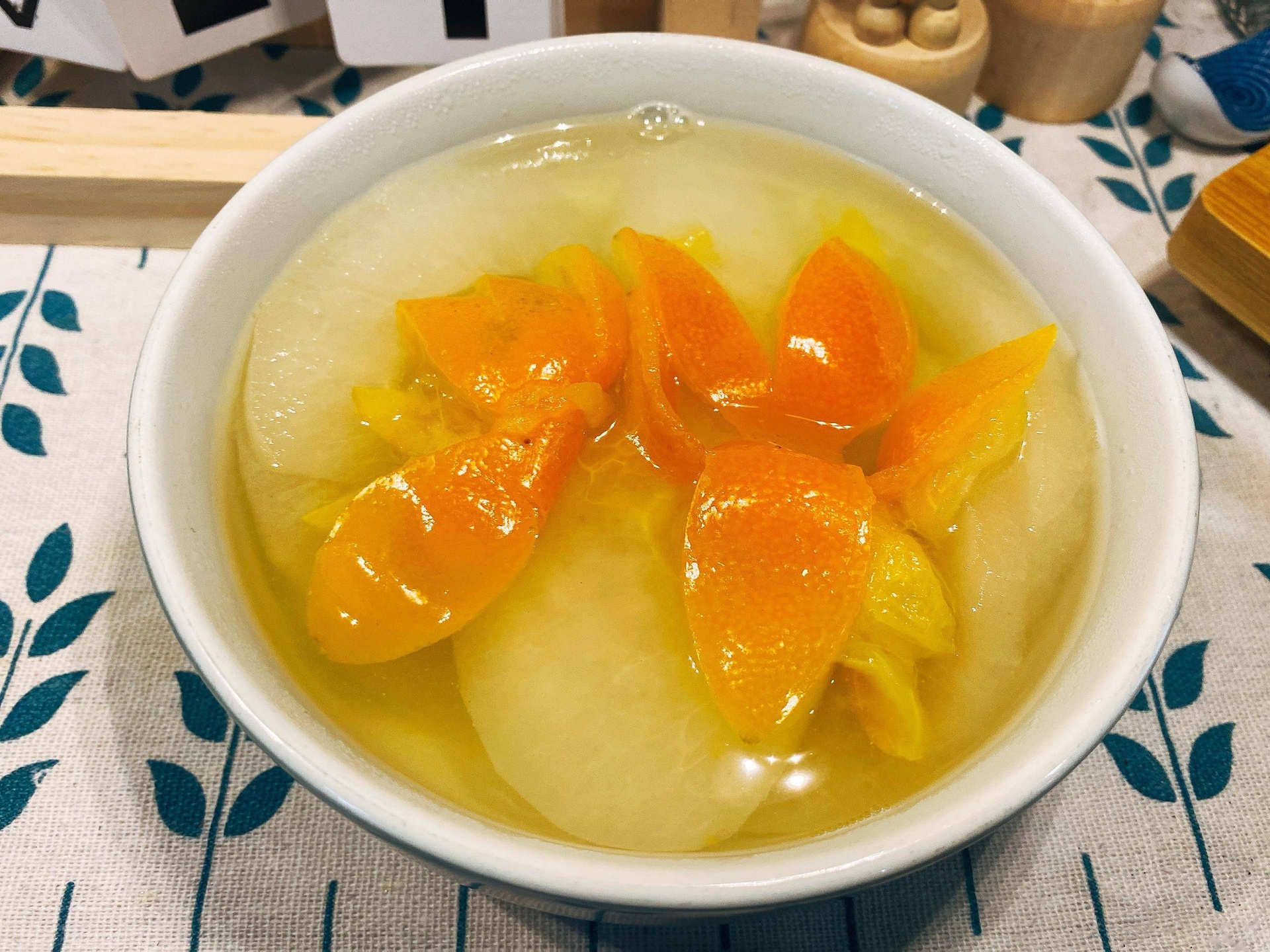 一碗刚出锅的甜橙炖盐盛放在白色陶瓷碗中,橙色果肉清晰可见,汤汁呈琥珀色,表面漂浮着几粒枸杞和一丝橙络,旁边配有木勺和透明玻璃杯,整体摆盘简洁温馨,背景为浅灰色桌布 甜橙炖盐成品展示