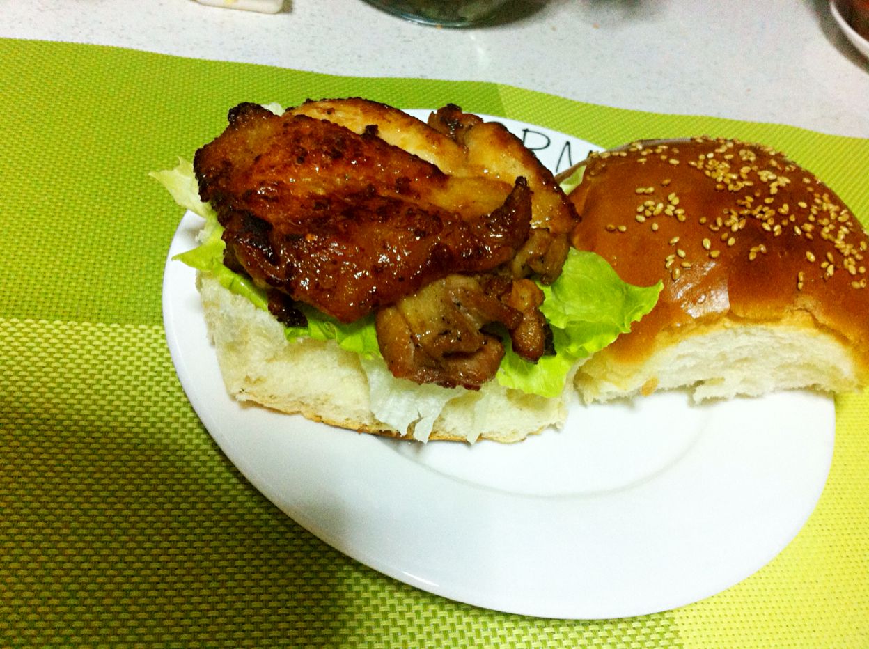 一个完整的照烧鸡腿堡被斜切展示,露出内部层次:底部为烤制微焦的汉堡胚,依次叠放生菜叶、琥珀色酱汁包裹的鸡腿肉、翠绿葱段,顶部为带芝麻的圆面包,旁边配有几根薯条和一小碟剩余照烧汁,整体摆盘精致,光线温暖,突出食物色泽与质感 照烧鸡腿堡成品展示