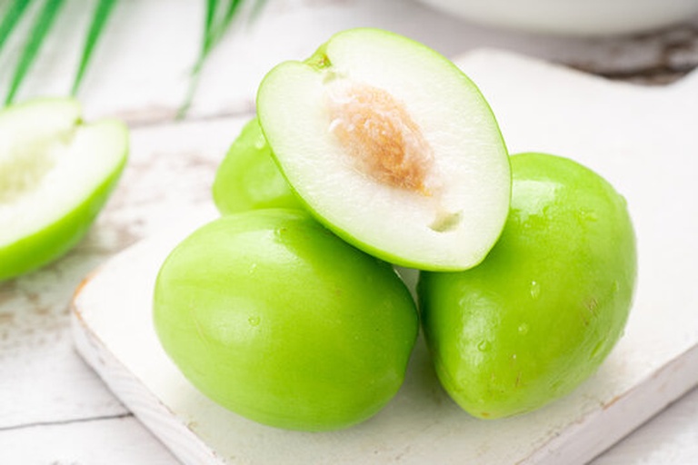 一颗新鲜青枣被横向切开,露出乳白色、水分饱满的果肉和中间较小的褐色果核,果皮呈鲜亮的翠绿色,切口处有晶莹的汁液渗出,背景为木质砧板和自然光环境 青枣切面特写图