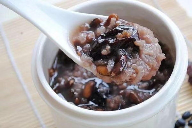 黑豆黑米杂粮粥成品图：深紫褐色浓稠粥体盛于青瓷碗中，表面浮有细密米油光泽，撒少量熟黑芝麻与枸杞点缀，勺子插入可见软糯黑米粒与完整黑豆嵌合状态