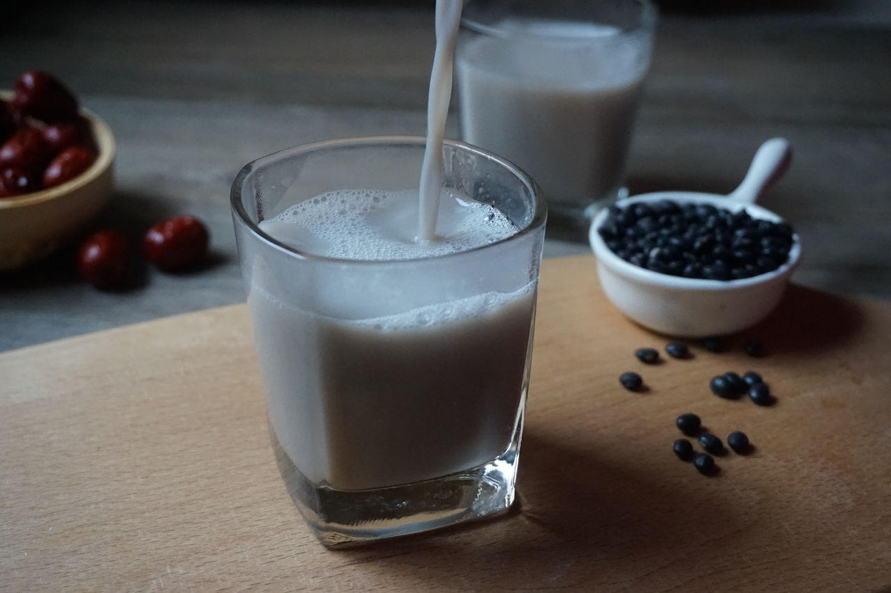 一只透明玻璃杯盛满深棕色略带光泽的黑豆豆浆,液体表面光滑无明显泡沫,杯子旁放着一碟泡发前的干黑豆作为对比,背景为木质餐桌和自然光线,突出食物的真实质感与健康属性 一杯成品黑豆豆浆特写