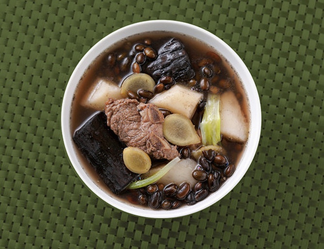 一碗热气腾腾的黑豆炖排骨,汤色浓郁,黑豆软糯,搭配绿色香菜点缀,盛放在白色瓷碗中,背景为厨房木质台面,体现家常健康饮食氛围 黑豆料理场景