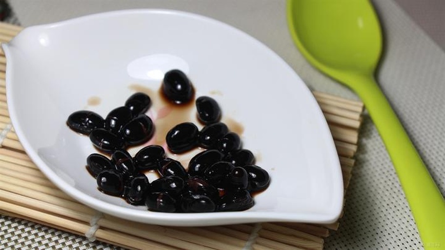 一位亚洲中年女性身穿浅色围裙,在明亮现代厨房中用透明玻璃锅煮黑豆水,锅中水面泛起紫红色泡沫,旁边摆放着量勺和干黑豆,窗台有绿植,整体画面传达健康生活理念 一位中年女性在厨房用玻璃壶煮黑豆水的场景