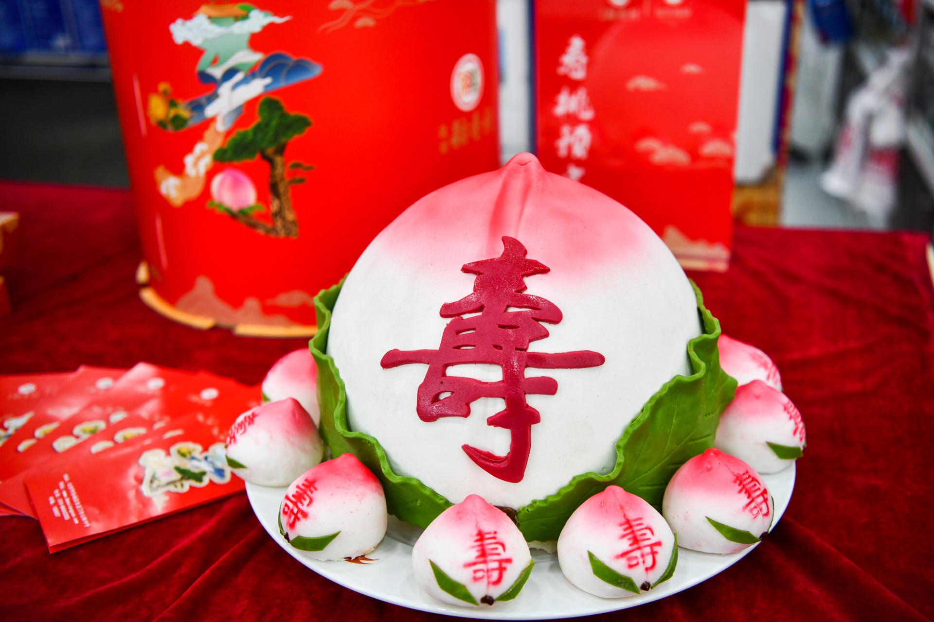 一个高达80厘米的巨大白色寿桃被安置在一个红色装饰底座上,顶部染成自然的粉红色,周围点缀着金色‘寿’字贴纸和小型花卉,背景为传统中式厅堂,灯光柔和,充满节日气氛 百斤面蒸寿桃成品全景展示