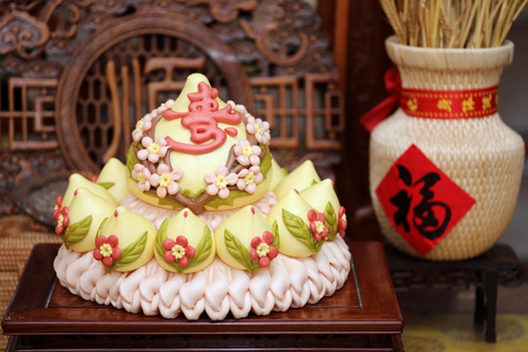 传统蟠桃主题民俗场景：古风庭院中，白发老者身着绛红唐装端坐主位，面前朱漆案几上陈列三枚硕大鲜红蟠桃与铜制寿字香炉，两侧孩童手捧桃枝与“寿比南山”卷轴，背景窗棂透出桃花剪影