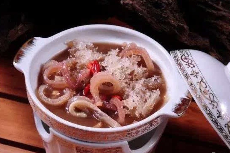 高清俯拍图:白色陶瓷炖盅盛装琥珀色清汤,汤面油光细腻,可见半透明鱼胶块与淡黄色生晒参片交错分布,旁边摆放银勺,背景为简约中式餐桌,营造温馨养生氛围 鱼胶炖洋参成品汤品展示